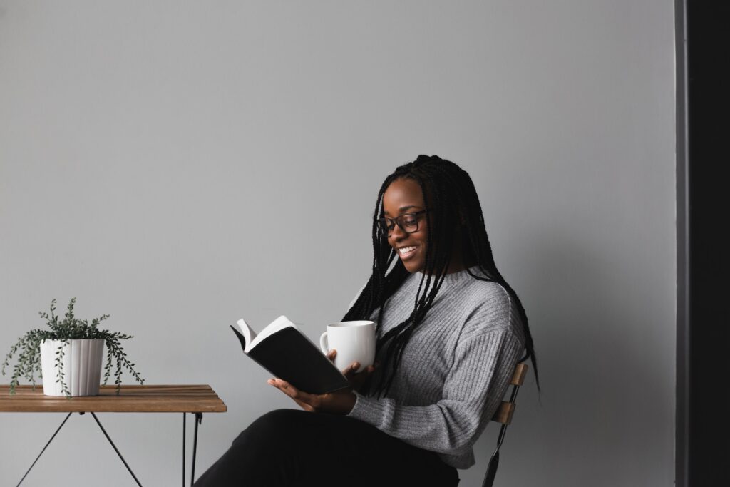 Woman smiling and reading a book with a black cover while holding a cup in another hand.